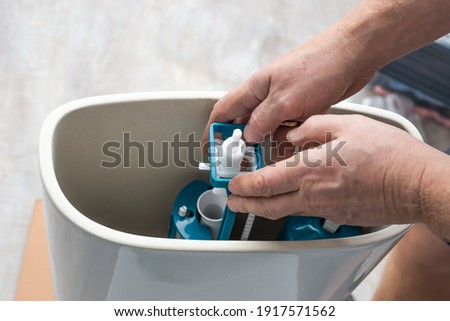 A plumber installs a water pump in a ceramic toilet cistern. Drainage system installation, home repair. Royalty-Free Stock Photo #1917571562