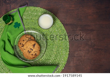 St Patrick's Day treat of cookies and milk on a glass plate with placemat, napkin, shamrock and tiny leprechaun hat on a wood table background