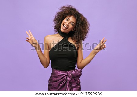 Optimistic lady in black top and purple skirt smiles on isolated background. Cool girl with curly hair shows peace sign on lilac backdrop..