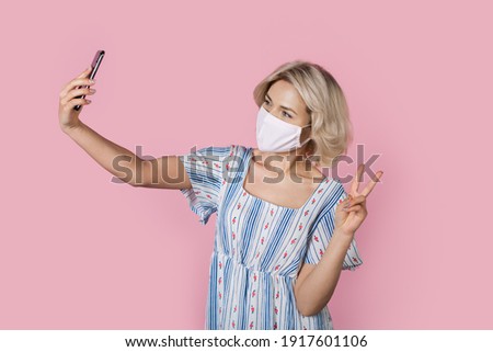 Adorable blonde lady in medical mask in gesturing the peace sign while taking a selfie on a pink wall
