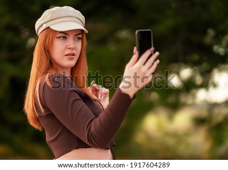 A young Caucasian red-haired woman in a hat doing a selfie in the park