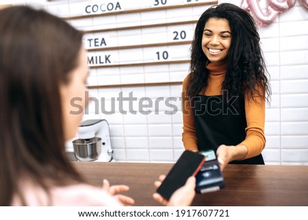 Cashless payment concept. Female visitor paying for order using modern technology, friendly black waitress give card reader machine for customer make payment transaction using cellphone