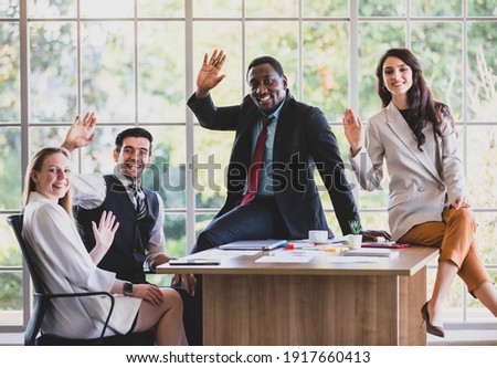 Group of four middle-aged African and Caucasian businessmen and businesswomen gathered around modern office working table, raising hands to agree in the meeting room while looking at the camera