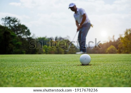 A picture of a golf ball about to pit. A blurred background of a golfer doing a golf putt. Beautiful golf course, evening time, the sun is setting