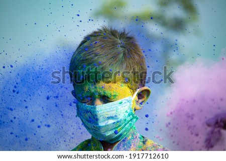 Portrait shot of young kid with face mask standing while colorful Holi powder throwing at him - concept of holi festival during coronavirus coivd-19 pandemic. Royalty-Free Stock Photo #1917712610
