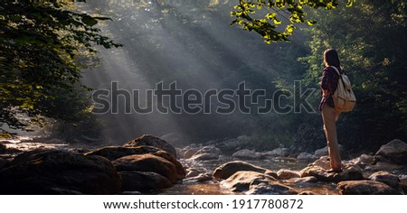 A woman explores new, magical, and fantastic places around the world, surrounded by nature and spreading her arms to breathe and relax. Female hiker crossing the forest creek. Royalty-Free Stock Photo #1917780872