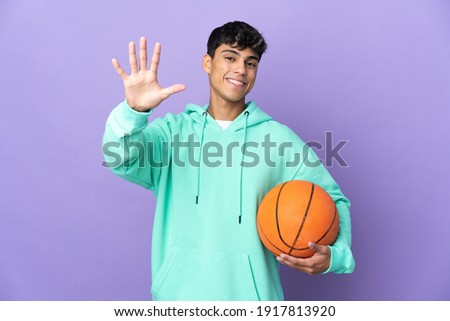 Young man playing basketball over isolated purple background counting five with fingers