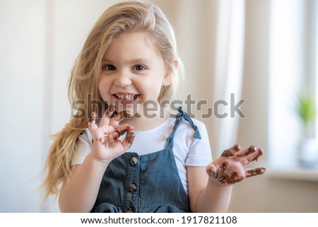 Portrait of adorable blonde little girl eating chocolate. Close up of toddler child with dirty funny face and fingers. . High quality photo Royalty-Free Stock Photo #1917821108