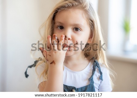 little blonde cute girl got her chocolate fingers dirty and licks them. High quality photo Royalty-Free Stock Photo #1917821117
