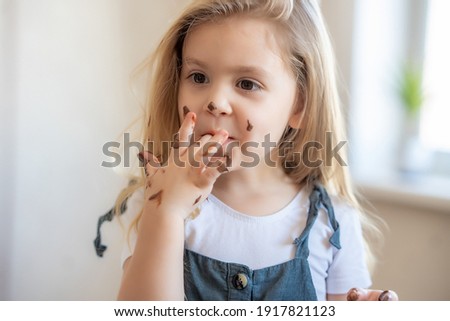 little blonde cute girl got her chocolate fingers dirty and licks them. High quality photo Royalty-Free Stock Photo #1917821123