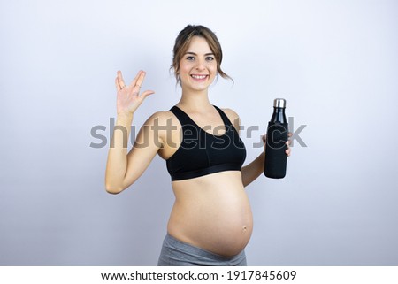 Young sportswoman pregnant wearing sportswear holding bottle with water over white background doing hand symbol