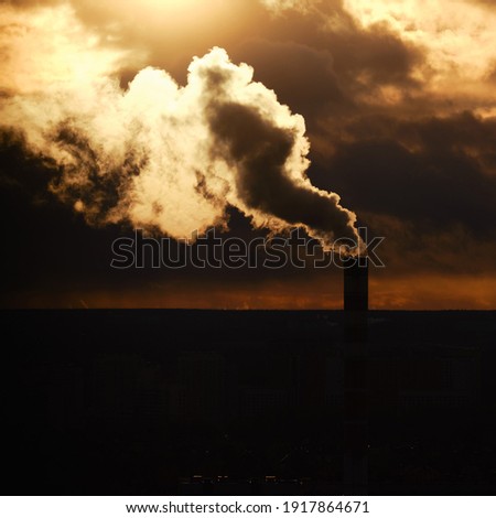 Silhouette of a chimney with white smoke coming over the city, evening sunset