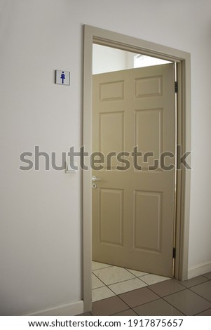 A light-colored open door to the women's toilet with a sign on the wall. The floor is covered with tiles. Frame in perspective