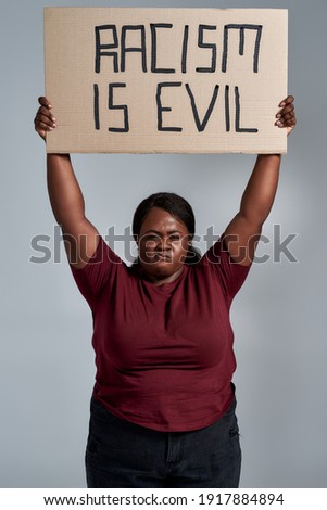 Fierce plus size young african american woman in casual clothes looking at camera, holding Racism is evil banner above her head, posing isolated over gray background. Social issues, protest concept