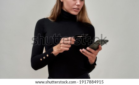 Cropped shot of caucasian young woman wearing black clothes holding credit card, using smartphone while posing isolated over gray background. Fashion, shopping, banking, payment concept. Web Banner