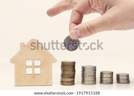 The hand adds a coin to the pile, there is a house nearby. Toning. The concept is the accumulation of money for housing, mortgages, affordable housing, the dream of owning a home, loan installments. Royalty-Free Stock Photo #1917915188
