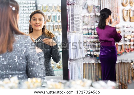 Smiling young woman shopping bijouterie, trying on necklace and looking at mirror