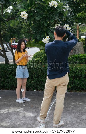 Male photographer take a photo of teenage model girl at the plumeria tree while she hold the flower and camera.