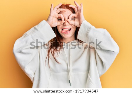 Young red head girl wearing casual sweatshirt doing ok gesture like binoculars sticking tongue out, eyes looking through fingers. crazy expression. 