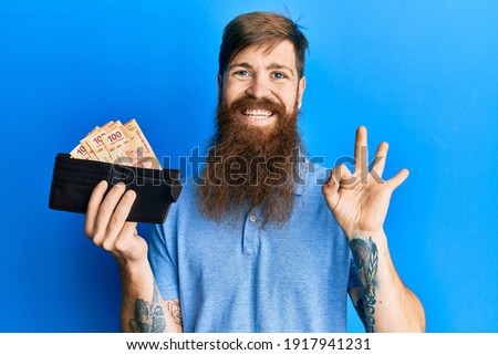 Redhead man with long beard holding wallet with mexican pesos doing ok sign with fingers, smiling friendly gesturing excellent symbol 
