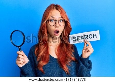 Young redhead woman holding magnifying glass and search word afraid and shocked with surprise and amazed expression, fear and excited face. 