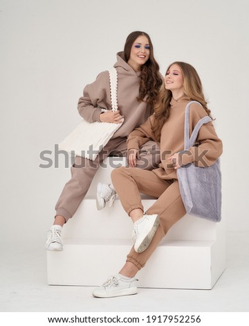 Two young female teenagers sitting at white stairs against white background. Pretty girl in sporty costumes trousers and hoodie holding textile bags