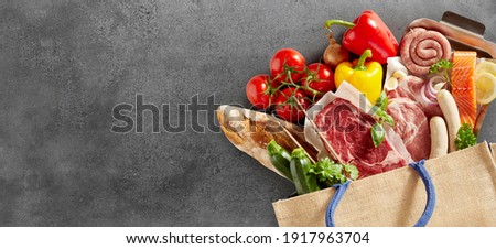 Close up on fresh groceries in a shopping bag over a textured slate background fresh groceries including assorted meat, fish, baguette, tomatoes, sweet peppers and herbs