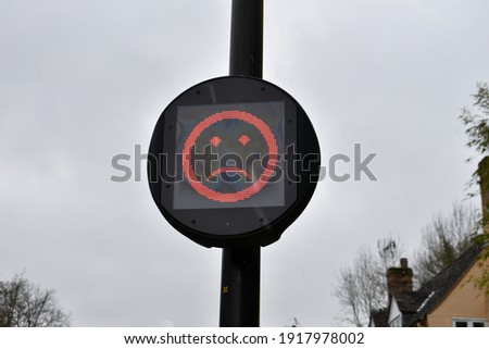 close up of red unhappy emoji face on a round traffic calming speed advisory sign outside on a black post on a grey rainy day