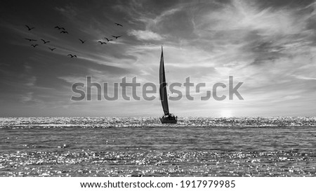 Black and white photograph of a sailboat sailing towards the horizon at sunset