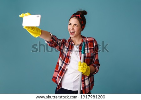 Excited beautiful girl in gloves with mop taking selfie on cellphone isolated over blue background