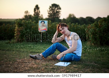 Young male artist, wearing torn jeans and white t-shirt, sitting on ground on green field during sunset, leaning on his hand, thinking. Painting workshop. Artistic inspiration concept.