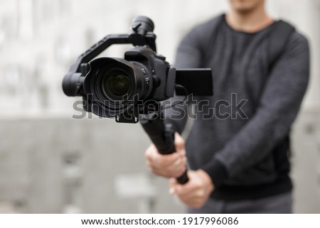 filmmaking, videography, hobby and creativity concept - close up of modern dslr camera on 3-axis gimbal in male hands Royalty-Free Stock Photo #1917996086
