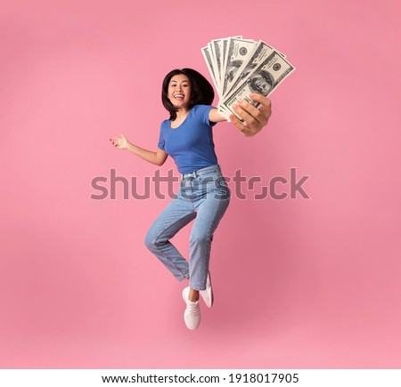 Full length portrait of a joyful young asian woman jumping up high and holding bunch of money banknotes, showing close to camera, celebrating win, isolated over pink studio background, collage Royalty-Free Stock Photo #1918017905