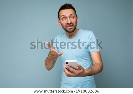 Photo of angry handsome young man with beard wearing everyday blue t-shirt isolated over blue background holding and using mobile phone communication online on the internet looking at camera and