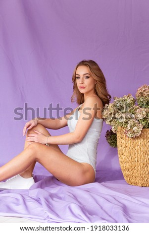 Beautiful woman in a white bodysuit on a purple background with a huge basket of flowers on the background. A slender girl on a purple background.