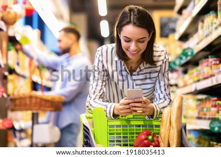 Smiling Female Customer Doing Grocery Shopping Using Smartphone Walking With Cart In Supermarket. Selective Focus. Woman Using Groceries Shopping Application On Phone Bying Food In Super Market Royalty-Free Stock Photo #1918035413
