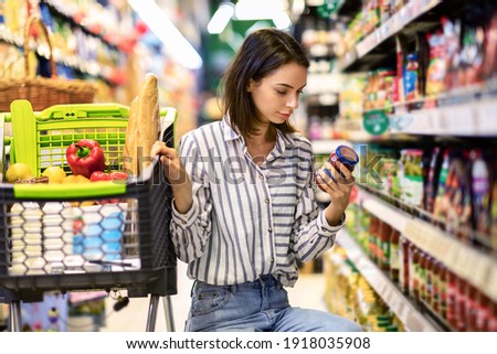 Consumption And Consumerism. Portrait Of Young Woman With Shopping Cart In Market Buying Groceries Food Taking Products From Shelves In Store, Holding Glass Jar Of Sauce, Checking Label Or Expiry Date Royalty-Free Stock Photo #1918035908