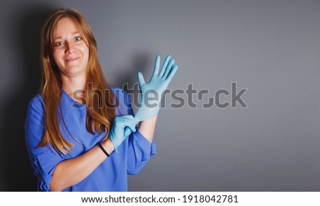 Advertising concept cosmetic and medical protection. Elderly female in blue lab coat puts on hands protective sterile gloves on gray background, protecting against bacteria and chemicals. Copy space