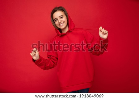 Photo shot of beautiful happy smiling brunette little girl wearing trendy red hoodie standing isolated over red background wall looking at camera