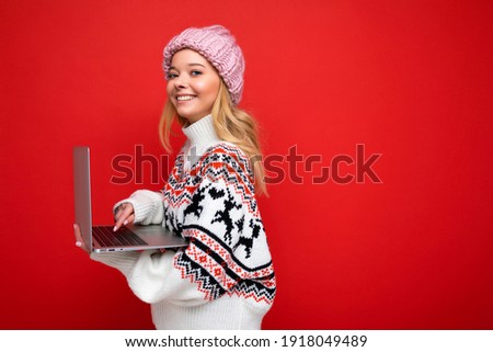 side profile photo of charming smiling happy pretty young woman holding laptop isolated over red wall background looking camera wearing winter hat and knitted new year sweater typing on keyboard