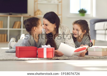 Cute family celebrating Mother's Day at home. Happy young woman lying on floor with her children, reading greeting card, thanking kids for present, touching noses and Eskimo kissing with her daughters
