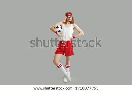 Full body happy satisfied nerdy man in glasses, headband, white tank top and red sports shorts standing akimbo, holding soccer ball and smiling isolated on grey background. Humorous football concept