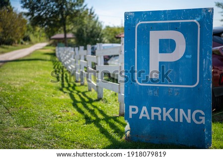 Big blue parking sign, leaning against old wooden white cattle fence, on a countryside road. A farm house in background.