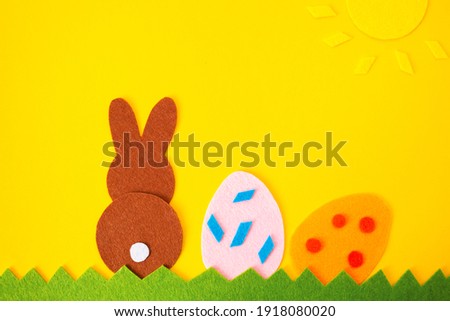 Easter holiday decorations. Cut out of felt applications of two eggs and brown rabbit on the grass with sun. Yellow background. Copy space. Flat lay.