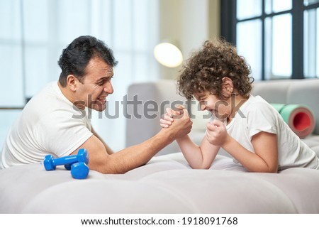 Cute latin boy trying to win while having fun, playing armwrestling together with his father at home lying on the couch