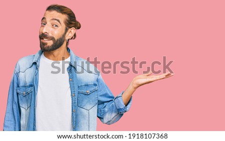 Attractive man with long hair and beard wearing casual denim jacket smiling cheerful presenting and pointing with palm of hand looking at the camera. 