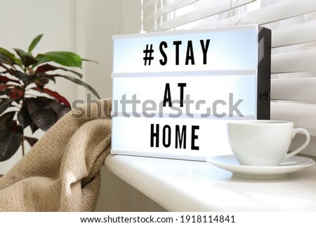 Cup of drink and lightbox with hashtag STAY AT HOME on window sill indoors. Message to promote self-isolation during COVID‑19 pandemic