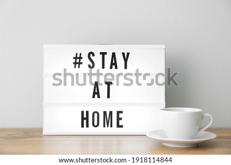Cup and lightbox with hashtag STAY AT HOME on wooden table near white wall. Message to promote self-isolation during COVID?19 pandemic