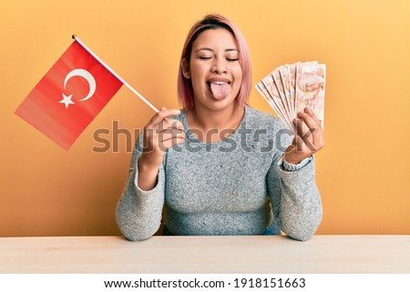 Hispanic woman with pink hair holding turkey flag and liras banknotes sticking tongue out happy with funny expression. 