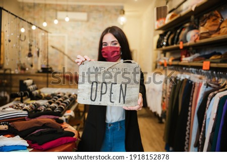 Portrait of a beautiful young clothing store sales assistant holding the "Welcome we're open" sign at the business entrance wearing a face mask during the Coronavirus Covid-19 pandemic